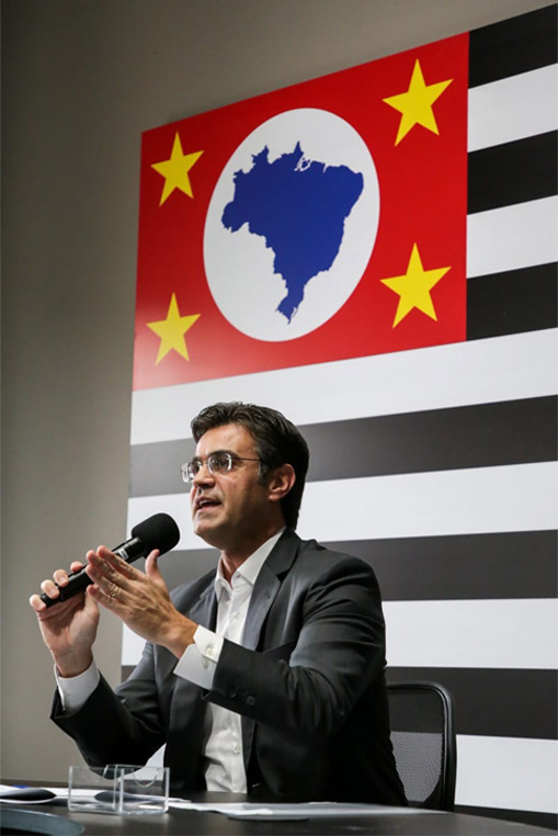 Descrição de imagem: fotografia colorida. Rodrigo está sentado, ele veste terno cinza escuro e camisa branca. Segura microfone com a mão direita e gesticula com a esquerda. Ao fundo, parede com quadro de bandeira do estado de SP. Fim da descrição.