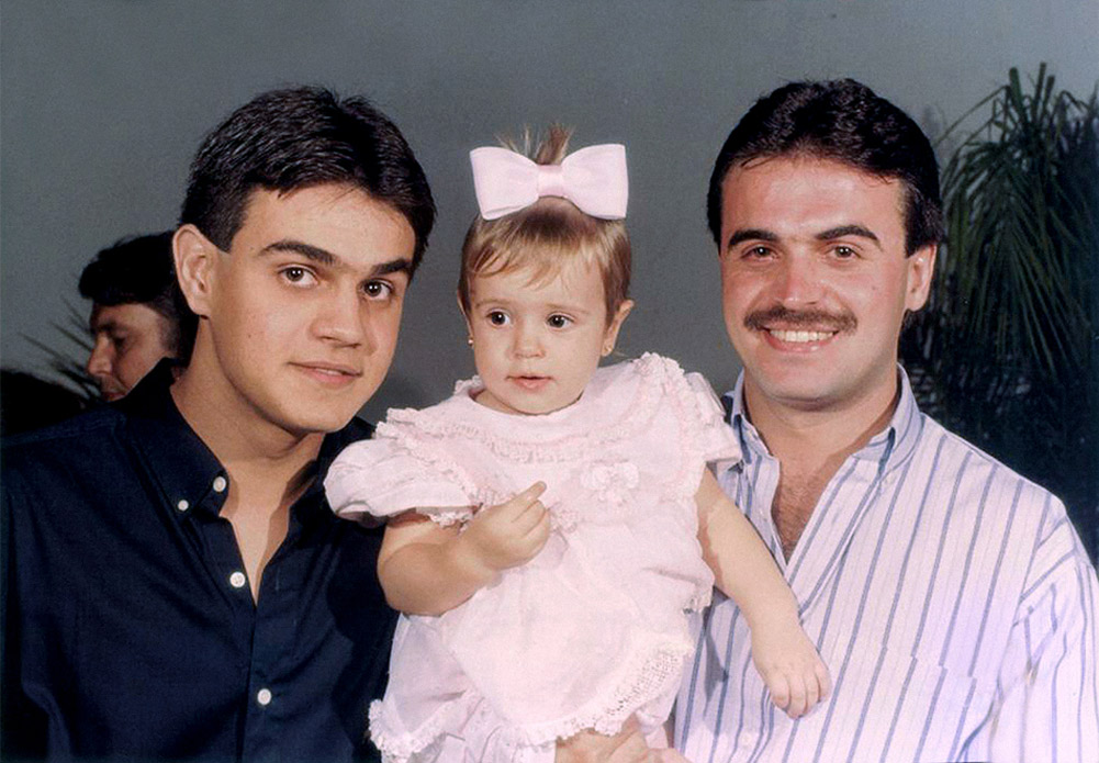 Descrição de imagem: fotografia colorida. Dois homens e um garoto em pé, fotografia tirada à luz do dia, em local com árvores. Na frente e em destaque estão João Pedro e Seu Paulino, filho e pai de Rodrigo, respectivamente. João Pedro é um garoto de pele branca e cabelo castanho claro, ele usa camiseta cinza com grafismos cinza escuro. Seu Paulino é um homem de pele branca e cabelo grisalho, ele usa chapéu branco, camisa azul e calça jeans. Rodrigo está ao fundo, ele sorri. Usa chapéu bege, camiseta marrom e calça jeans escura. Fim da descrição.